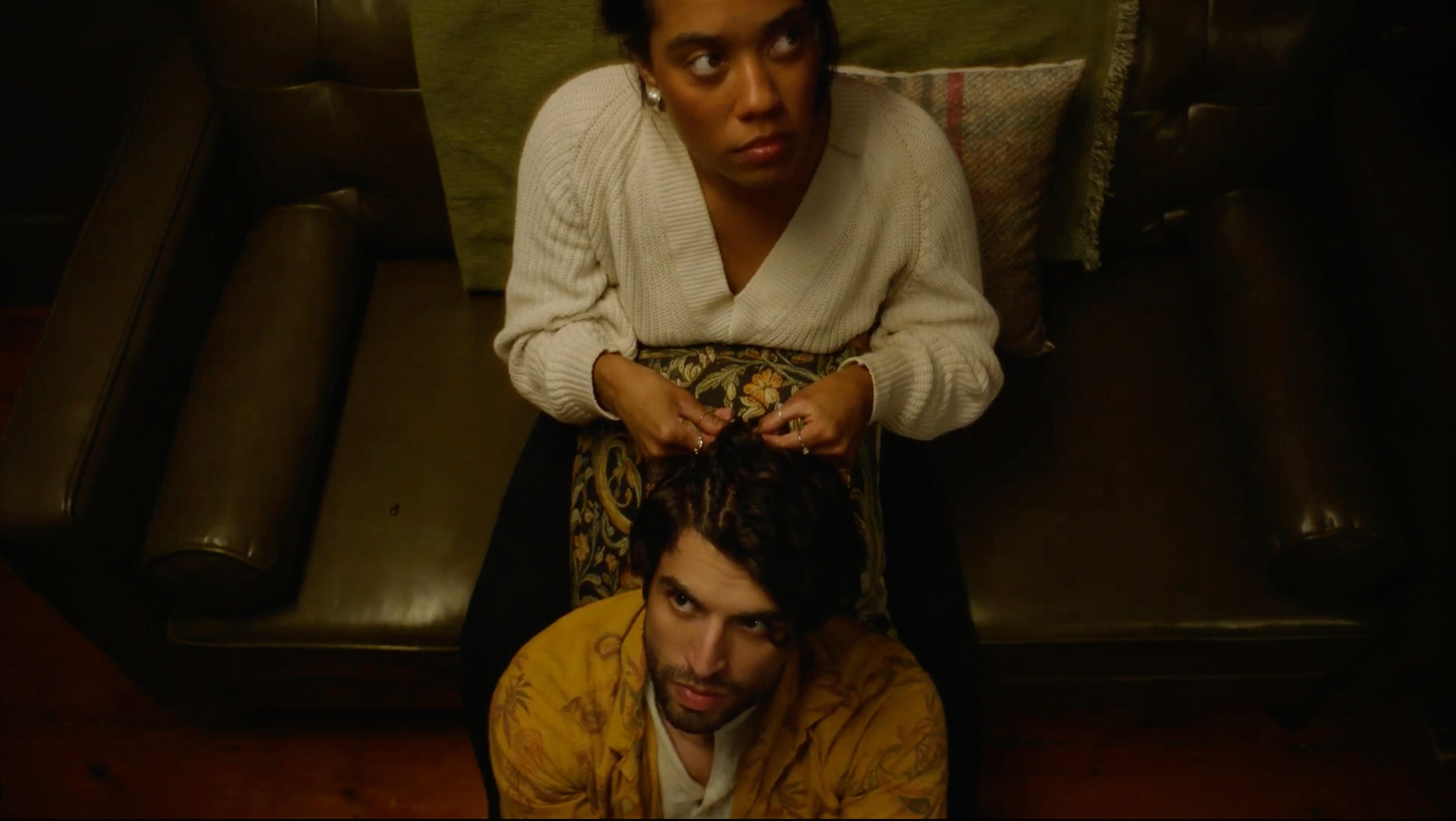 Woman on olive green couch braiding cornrows into hair of man sitting on floor.