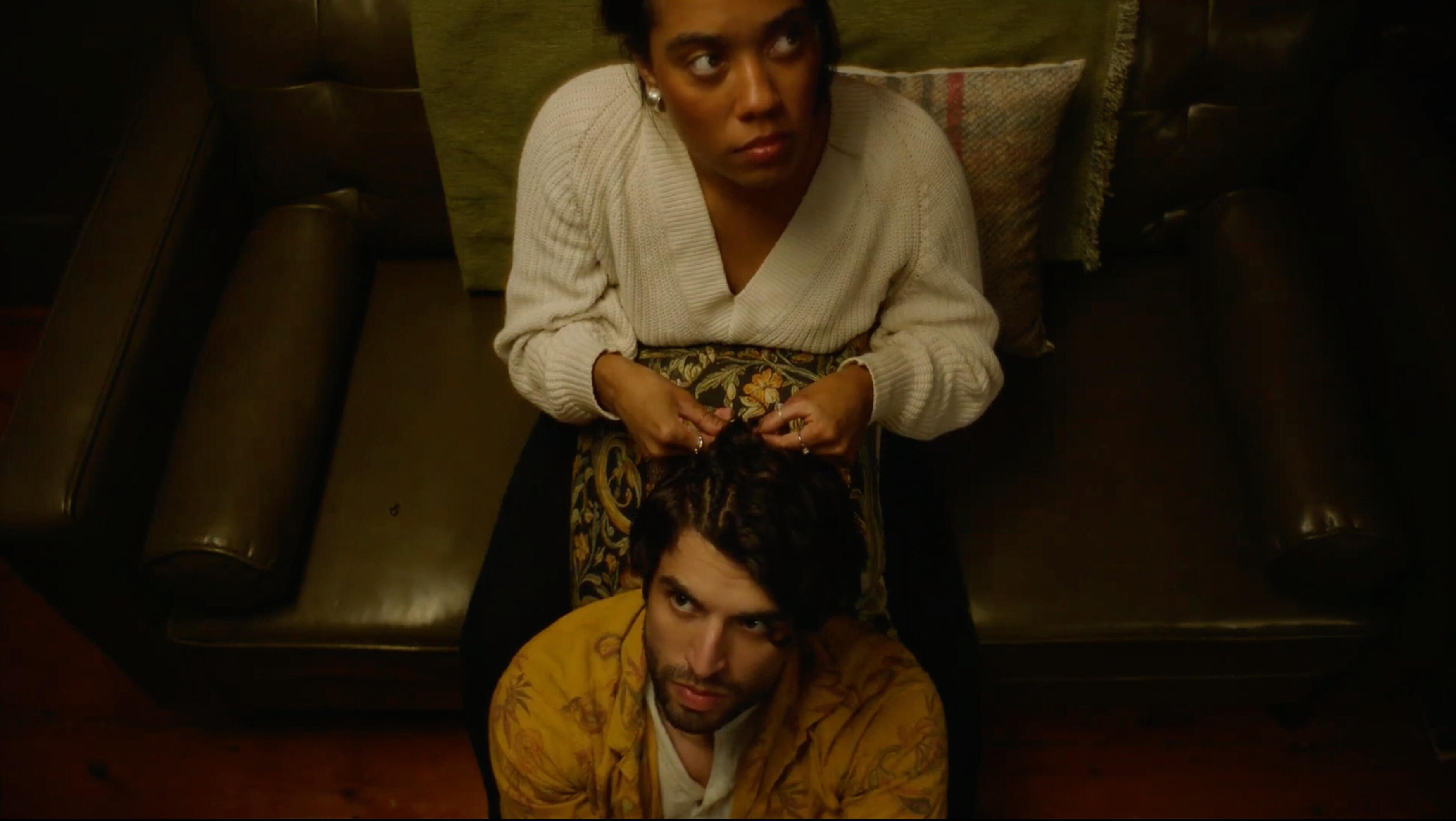 Woman on olive green couch braiding cornrows into hair of man sitting on floor.